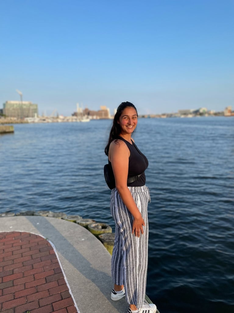 An image of Kaitlin. She is wearing her mid-length black her half-up half down and she is smiling at the camera. She is wearing a black tank top, striped grey and white linen pants, and white sneakers. She is standing by the Baltimore Harbor.